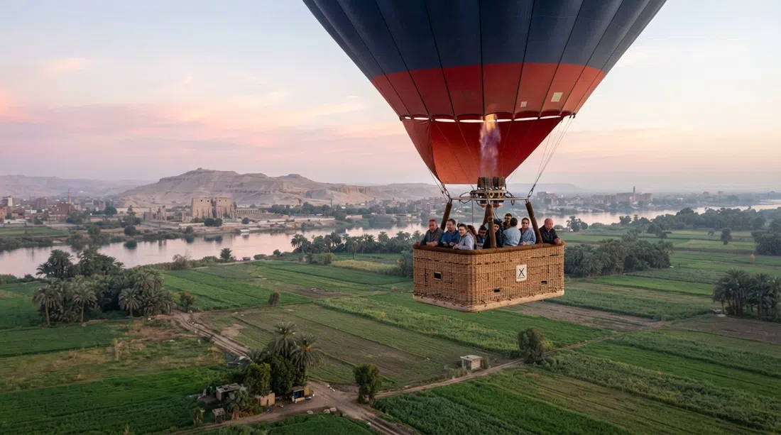 un globo aerostático al amanecer de Sindbad Balloons elevándose sobre los campos de la orilla oeste, con el Nilo y el paisaje del templo bajo un cielo rosa pálido